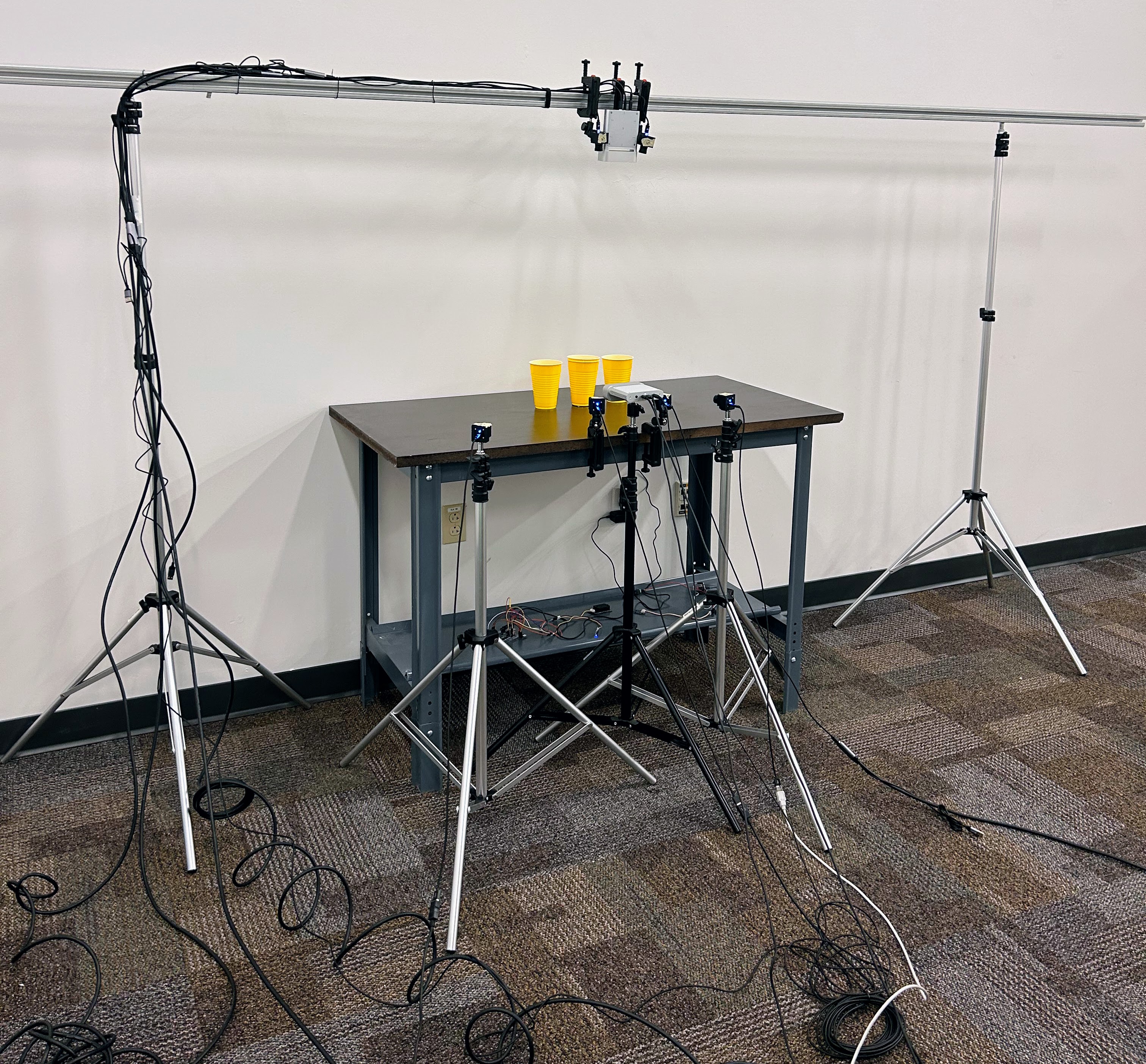 cameras in on tripods and racks are pointing at 3 plastic yellow cups on a table while lots of cords litter the ground